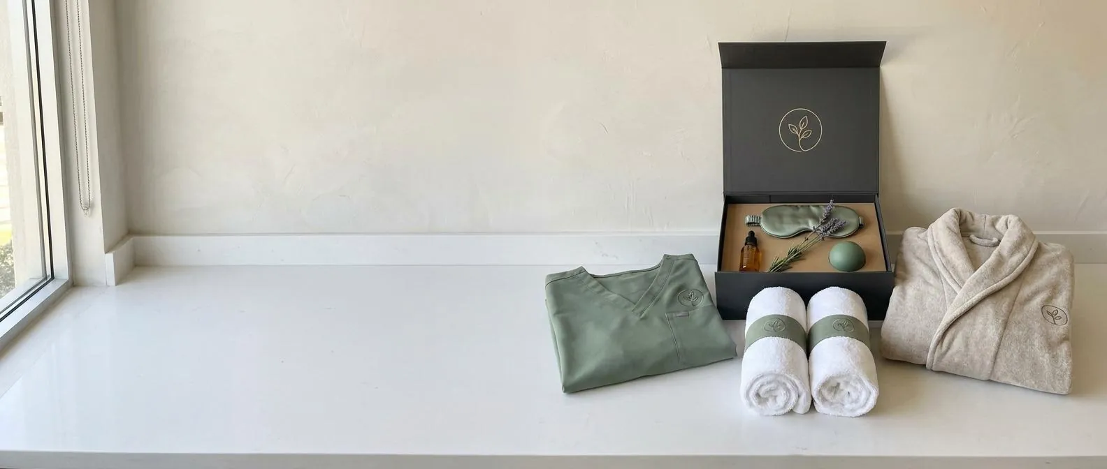 A bright med spa room with a white quartz counter running the full width, empty on the left; on the right, a presented kit with the same small sprig mark on every piece: folded sage scrubs, a plush oatmeal robe, two rolled white towels with sage bands and an open charcoal gift box holding a facial oil, a silk sleep mask, a stress ball and a sprig of lavender.