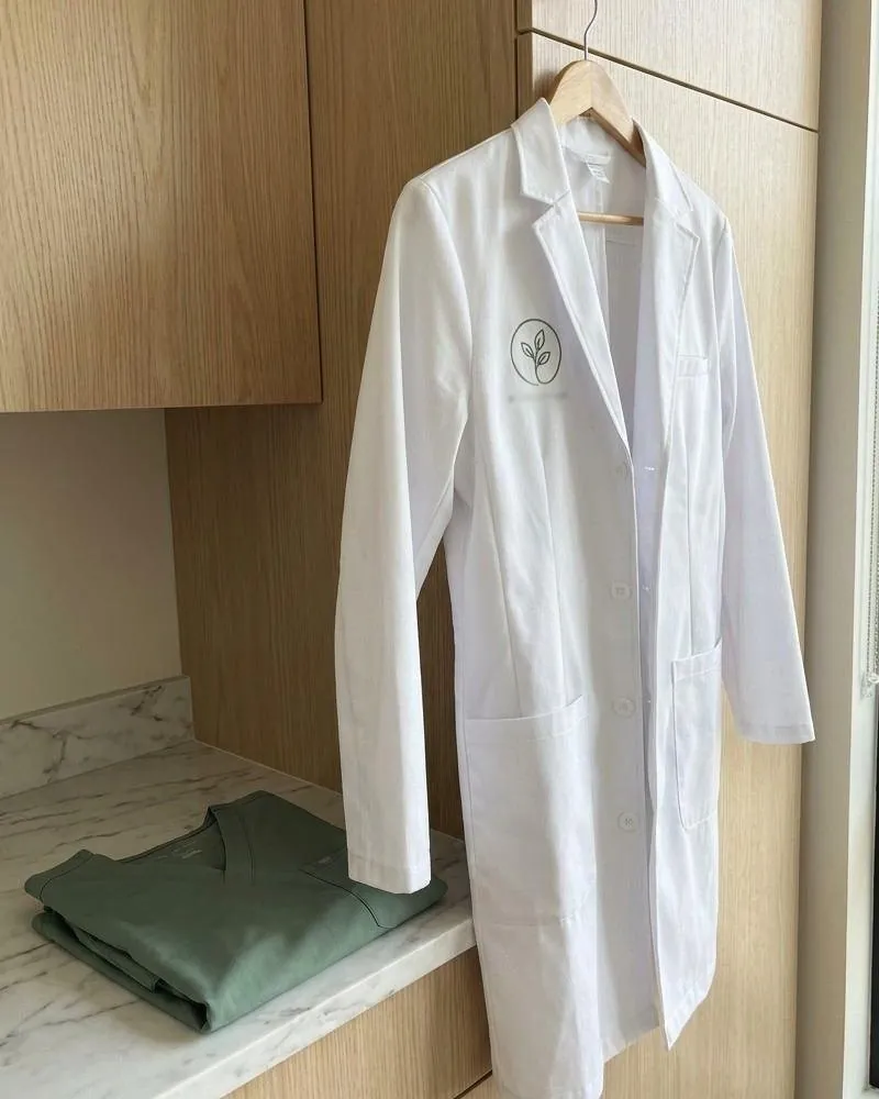 A tailored white lab coat with a sage embroidered logo on the chest, hanging above a folded scrub top.