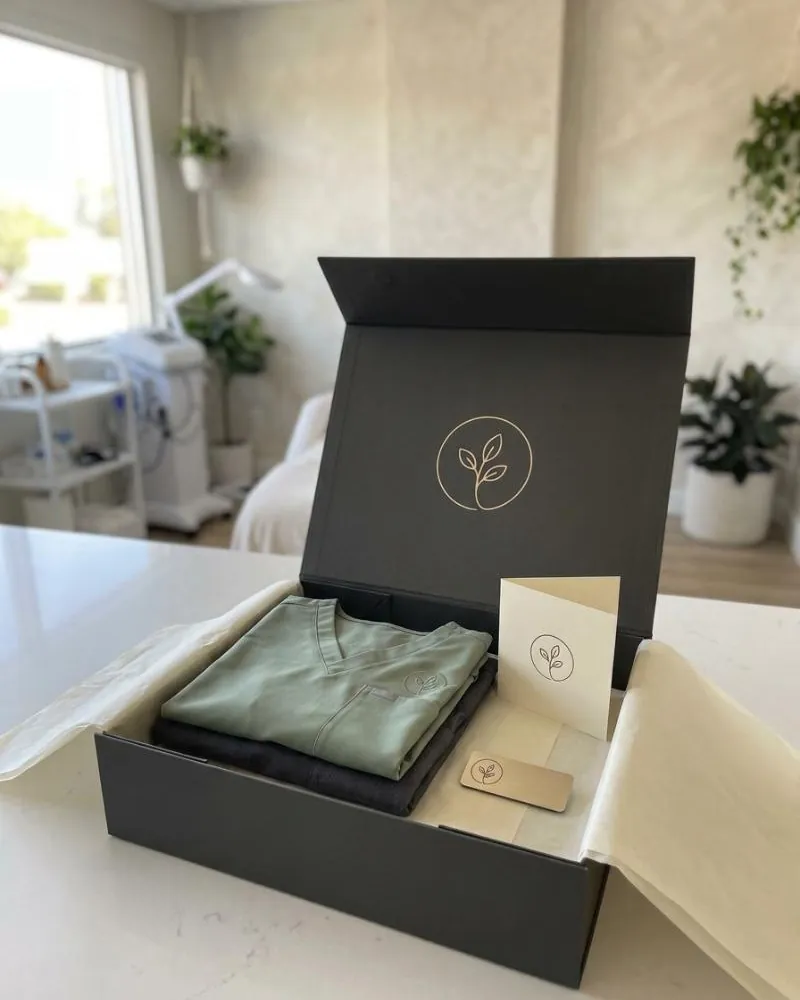 An open charcoal box on a treatment room counter holding two folded scrub tops, one sage and one charcoal, a champagne-gold name tag and a folded card, the mark foil-stamped inside the lid.