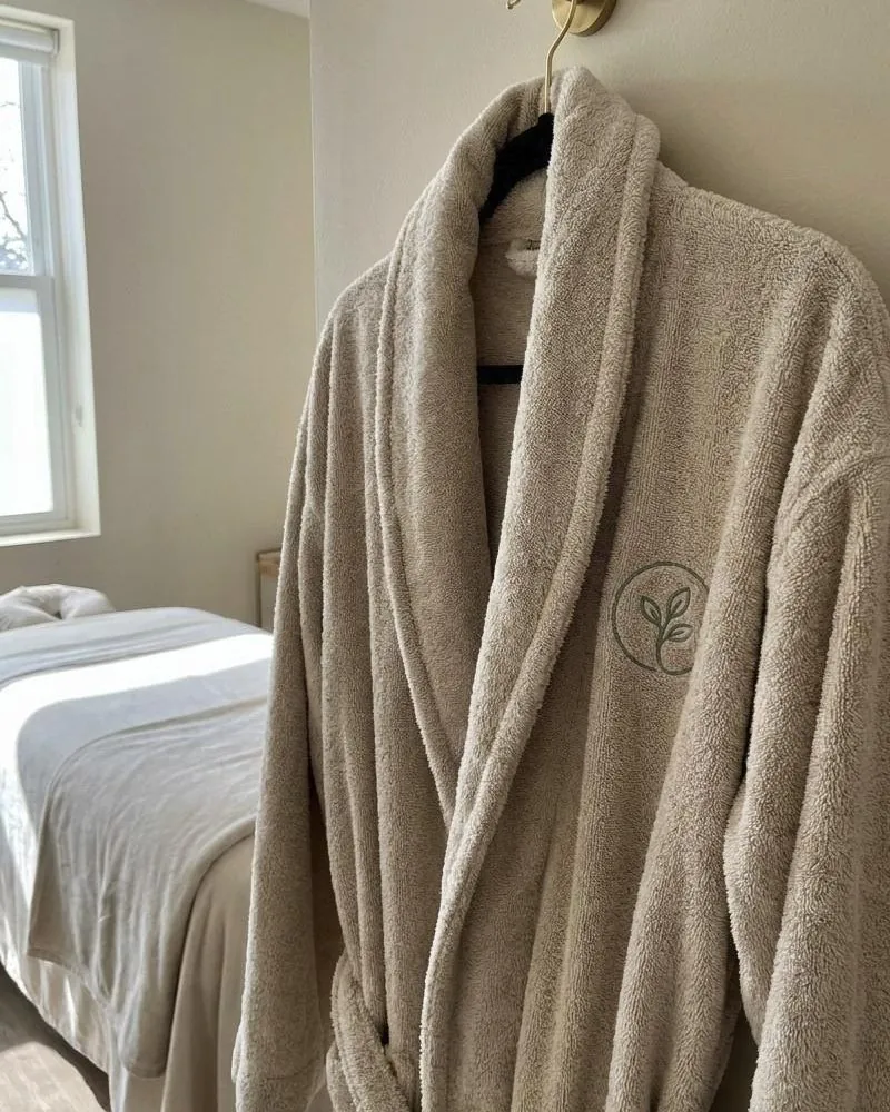 A plush oatmeal terry robe with a sage embroidered logo, hanging on a brass hook beside a treatment bed.