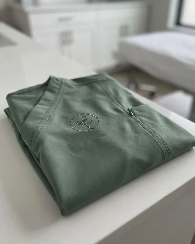 A folded sage scrub top in matte stretch fabric with a tonal embroidered logo on the chest.
