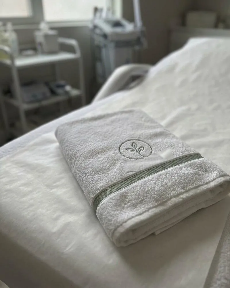 A folded white towel with a sage band and an embroidered logo, on a treatment bed.
